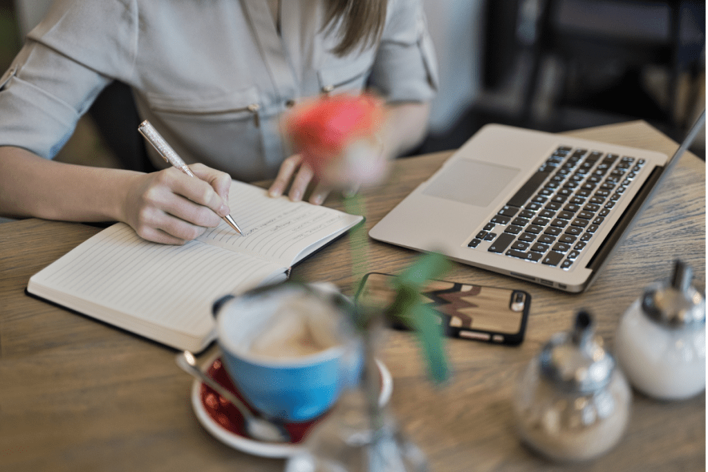 A woman sits at a table with a laptop and a notebook.