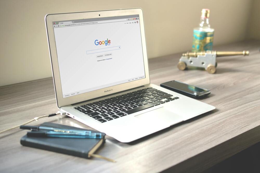 Laptop on a wooden desk with Google search open, alongside a smartphone, notebook, and decorative items.