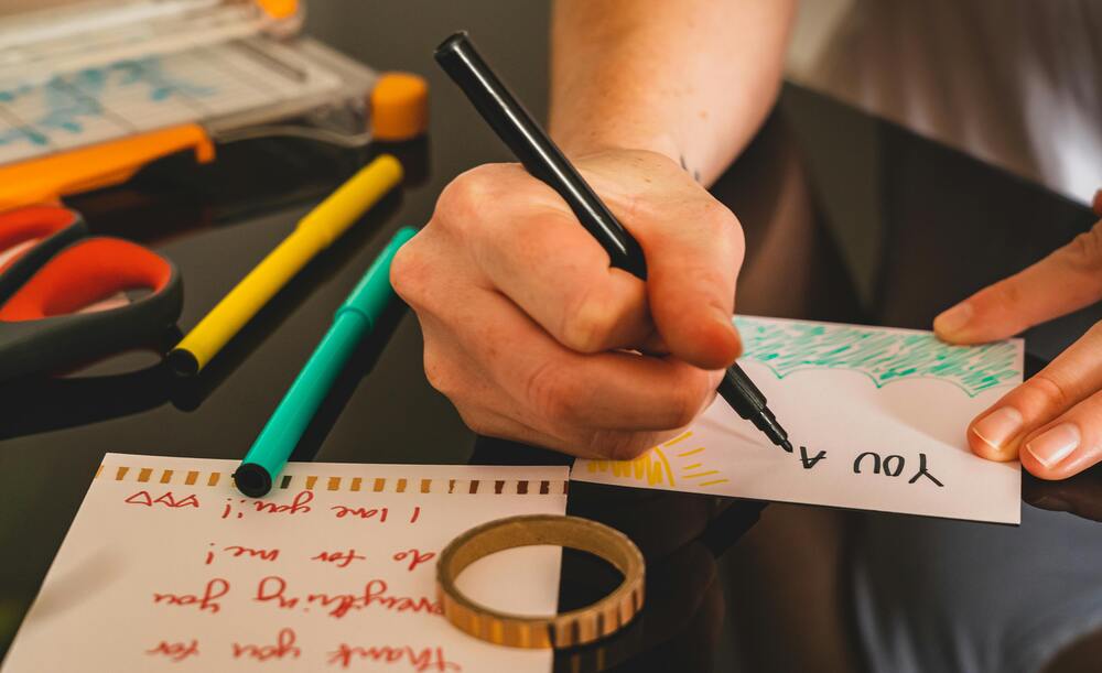 Person writing colorful handmade thank you card with markers and scissors.