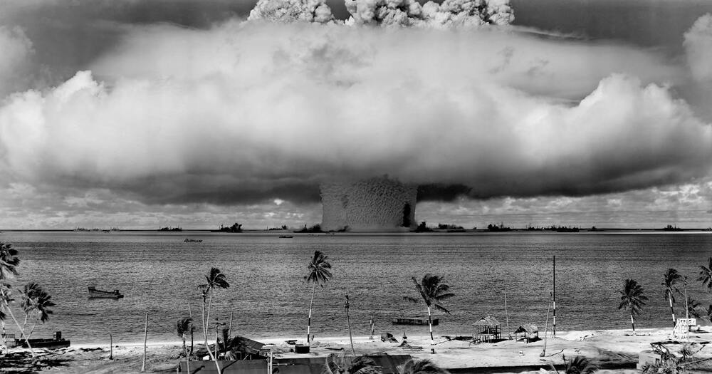 Mushroom cloud rising over ocean with palm trees and boats in the foreground.