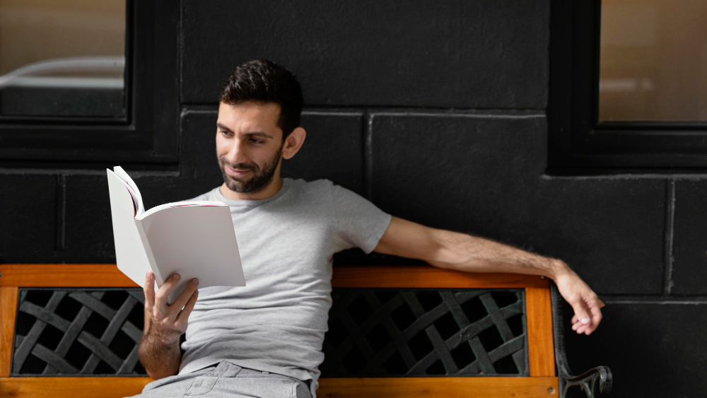 Man relaxing on a bench while reading a book in a peaceful setting against a dark wall.