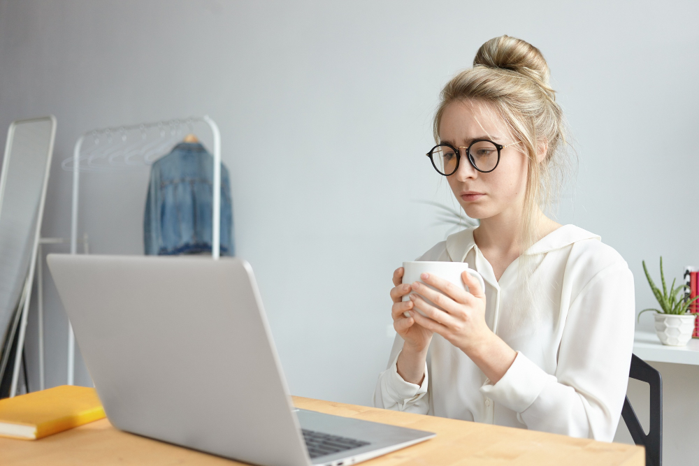 Woman in glasses working on a laptop at home office, holding a mug.